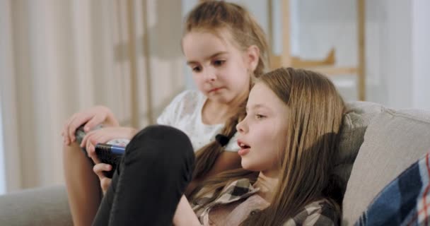Портрет двух харизматичных девушек, играющих вместе в игру, используя смартфон, сидя на диване, бабушка читает книгу рядом с ними и смотрит на своих внуков. 4k — стоковое видео