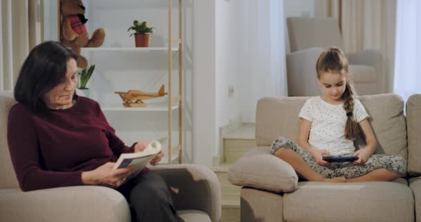 Carismática abuela mientras lee un libro en el sofá la sobrina jugando un juego en el teléfono inteligente sentado en el sofá . — Vídeos de Stock