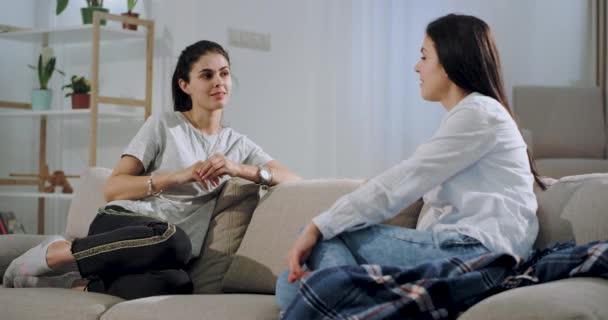 Dos amigas sonriendo y charlando muy amables en la sala de estar en el sofá, pasan un buen rato juntas . — Vídeos de Stock