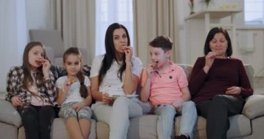 Geniş bir oturma odasında kanepede oturan kamera önünde bir Tv izlerken çocukları ve bisküvi yiyen bir büyükanne ile çok çekici anne