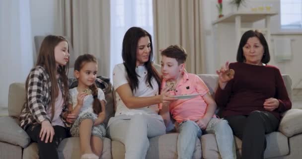 Molto felice e bella famiglia nipoti nonna e madre prendendo alcuni snack prima di iniziare a guardare un film di fronte alla fotocamera mentre seduto sul divano — Video Stock