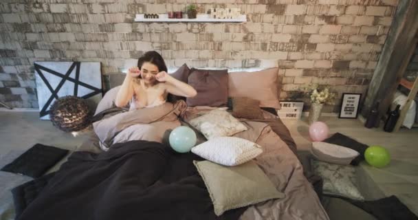 In modern loft bedroom charismatic lady waking up in the morning after a birthday party she stretching body with smile looking around, colorful balloons around — стоковое видео