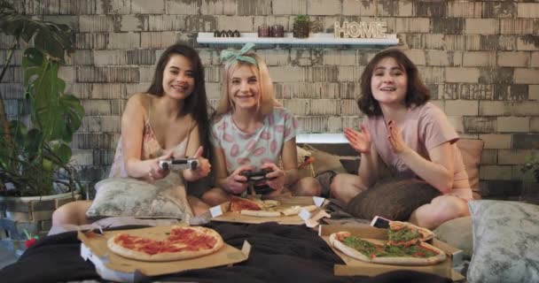 Три харизматичные дамы дома, в спальне, играют в игру PlayStation, едят пиццу и быстро проводят время вместе, надевая пижаму — стоковое видео