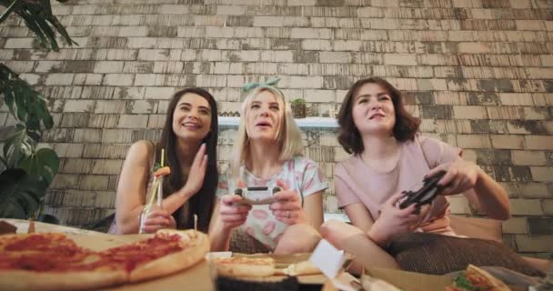 I ett modernt sovrum med en vacker design tre vänner damer spelar i PlayStation och äta pizza mycket koncentrerad och entusiastisk, klädd i en pyjamas, leende ansikten mycket attraktiv. — Stockvideo