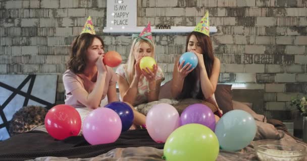 Carismático joven adolescente niñas soplando globos de colores delante de la cámara en la cama con un pijama, fiesta de cumpleaños celebrando . — Vídeo de stock