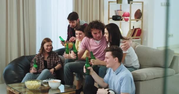 En un grupo multiétnico de amigos sentados en el sofá en una amplia sala de estar y viendo un partido de fútbol apoyan a su equipo favorito esperando un gol mientras beben oso y comen un poco de — Vídeos de Stock