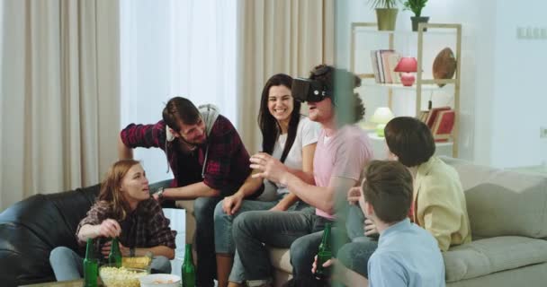 En una gran compañía de amigos multiétnicos disfrutando del tiempo juntos en una amplia sala de estar, uno de ellos jugando en un juego virtual usando gafas de realidad virtual — Vídeo de stock