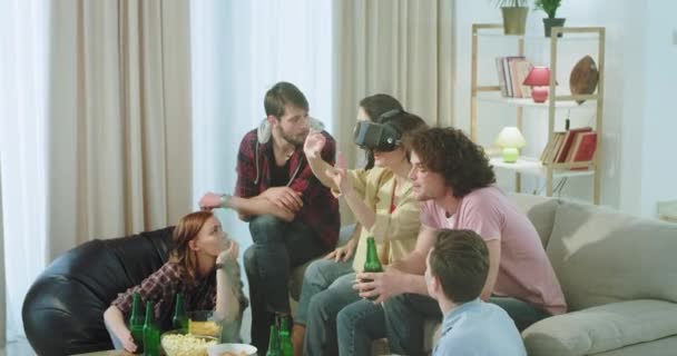 Una gran compañía de amigos multiétnicos en una sala de estar que tienen un rato divertido juntos, señora usando unas gafas de realidad virtual para explorar el mundo que están muy impresionados — Vídeo de stock