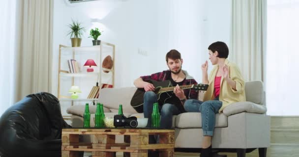 O doamnă drăguță doi prieteni cu păr scurt și un tip într-o cameră de zi se distrează împreună cântând la o chitară și cântând petrec un timp minunat împreună. — Videoclip de stoc