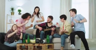 Arkadaşlar çok etnik Çekici büyük şirket oturma odasında birlikte iyi vakit geçirdik , bir gitar oynarken kanepede adam bazı , arkadaşlar dans ve komik hareket