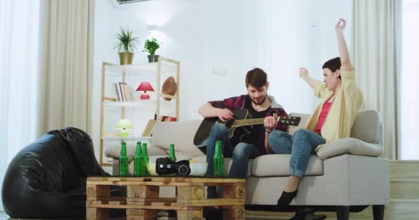 Nagyon karizmatikus hölgy egy srác énekel egy gitáron a kanapén egy tágas nappaliban ülve a kanapén — Stock videók
