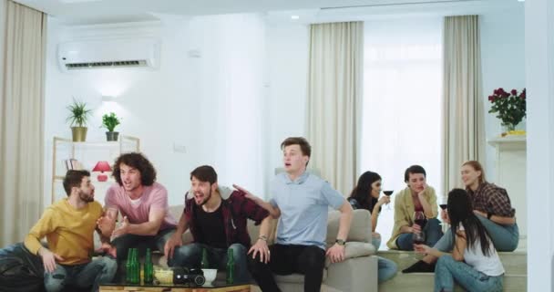 Attraktiva multietniska vänner killar och damer tittar på TV en fotbollsmatch de firar segern för sitt bästa lag i ett modernt hus damer dricker vin på bakgrunden — Stockvideo