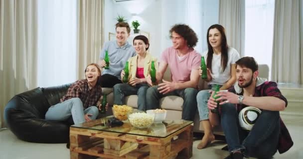 Bardzo podekscytowani przyjaciele przygotowują się do obejrzenia meczu piłki nożnej na telewizorze w środku salonu piją piwo i cieszą się razem — Wideo stockowe