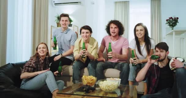 Attractive friends smiling and excited watching a football match on front of the TV they very enthusiastic yelling and clapping hands while their football team make a goal , they holding a bottles of — Stock Video