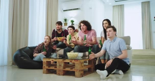 En una amplia sala de estar una gran compañía de amigos que disfrutan del tiempo mientras ven un partido de fútbol frente a la televisión están tan emocionados que su equipo es el primero y el mejor, cerveza y algunos — Vídeos de Stock