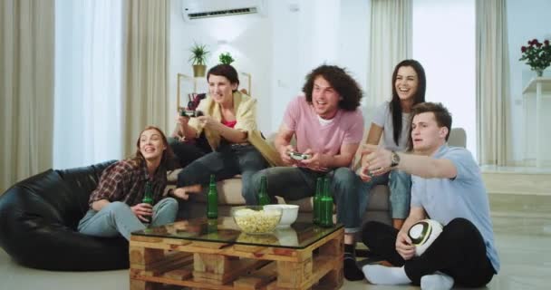 Nagyon karizmatikus fickó göndör haja és nagy mosoly játszik a PSP előtt a kamerát, miközben barátaik nézi, hogy a játék, nagyon izgatott sört iszik egy tágas nappaliban — Stock videók