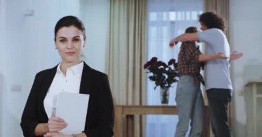 Kamera önünde güzel ev ajan kameraya bakarak ve arka planda onu tablet tutarak mutlu ve heyecanlı bir çift ajan ile büyük bir anlaşma yaptıktan sonra birbirlerine sarılma