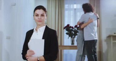 Büyük bir geniş oturma odasında büyük bir oturma odası awesome womna ev ajan siyah bir takım elbise li bir tablet holiding kameraya doğru bakarken, müşteri arka planda mutlu arka planda birbirlerine sarılma sonra
