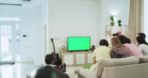 У сучасній просторій вітальні група друзів їсть деякі закуски і спілкується, сидячи на дивані і дивлячись фільм зелений екран по телевізору . — стокове відео