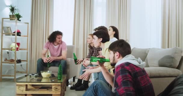 No sofá em uma espaçosa sala de estar, pessoas multi étnicas assistindo concentrado um jogo desportivo enquanto bebe cerveja e comer alguns lanches, eles apoiam sua equipe favorita com um rosto expressivo — Vídeo de Stock