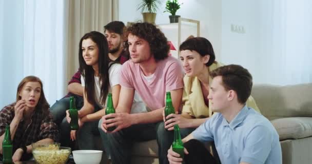 Spokojení a nadšené přátele, kteří sledují něco zajímavého v televizi popíjení piva a vychutnávají si čas spolu v prostorných obývacím pokoji na pohovce. — Stock video