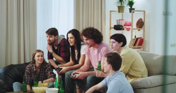 Muito carismático grande companhia de amigos estão muito animados aproveitar o tempo juntos, enquanto dois dos amigos que jogam em um jogo de vídeo — Vídeo de Stock