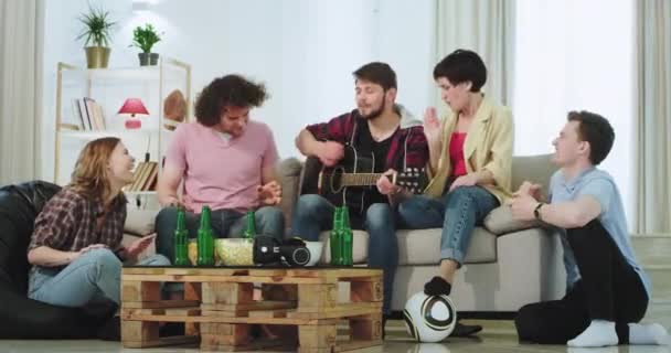 Egy modern, tágas nappali csoport baráti élvezni az időt együtt a kanapén játszik egy gitár énekét és boldog kiadások egy nagy idő
