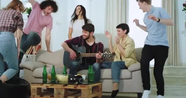 Una pareja tiene una fiesta en casa con sus amigos en la sala de estar cantando en una guitarra divertida dacing y sintiéndose tan emocionados y llenos de energía — Vídeo de stock