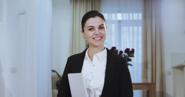 Перед камерой портрет привлекательной деловой женщины в костюме, улыбающейся большими белыми зубами и держащей планшет в руке — стоковое видео