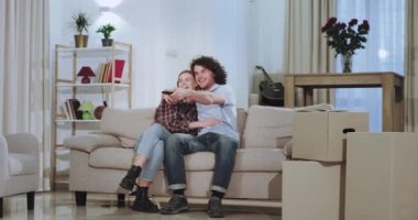 Gülümseyen ve karizmatik genç çift yeni bir evde zaman zevk , onlar tv önünde kanepede oturan hareketli bir günün ardından etrafında kutuları ile ve rahatlatıcı