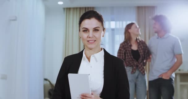 In een ruim huis een makelaar womna zeer mooi op zoek serieus naar de camera en het houden van een Tablet dan glimlach zeer groot met een perfecte tanden terwijl op de rug een paar erg opgewonden — Stockvideo