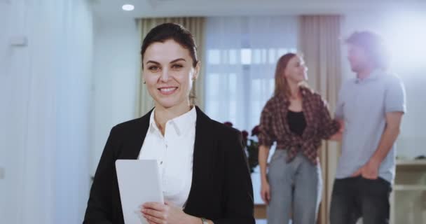 Улыбающиеся красивые женщины дома агента, смотрящие прямо в камеру, в то время как на заднем плане пара обнимаются счастливы друг с другом после подписания контракта — стоковое видео