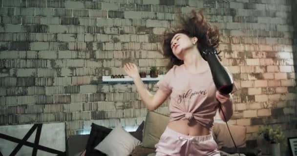 잠옷을 입은 카리스마 넘치는 아가씨가 헤어드라이어를 사용하여 아침에 카메라 앞에서 침대에서 신선하고 건조한 머리카락을 얻습니다. 4k — 비디오