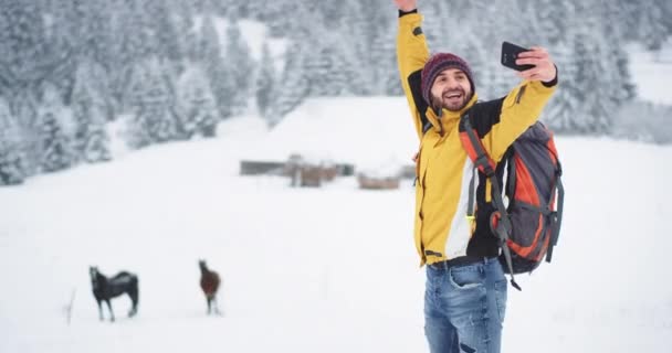 Lächelnd großer junger Mann Tourist, der in der Mitte der Berge an einem Wintertag unterwegs ist, kam er auf dem Land an und machte Selfies mit Pferden — Stockvideo