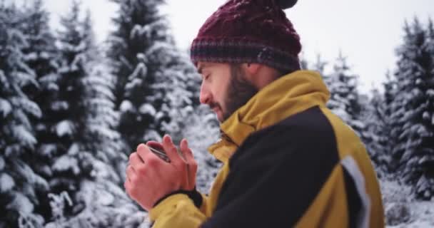 Хороший молодий хлопець посеред засніженого лісу захоплюється всією красою навколо, п'є гарячий напій з чашки заліза і зігрівається — стокове відео