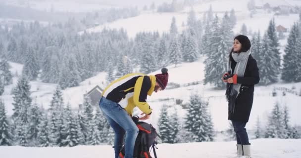 Twee vrienden toerist een man en vrouw, aangekomen in een geweldige plek in de top van het veld met sneeuw ze hebben een pauze te bewonderen alle schoonheid rond, besneeuwde bos, man nemen iets van zijn oranje — Stockvideo