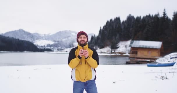 Charismatic jovem turista sorrindo grande na frente da câmera e segurando uma xícara de ferro com bebidas de chá para se aquecer ele de pé no lugar incrível com um grande lago e neve floresta e montanha, vestindo um — Vídeo de Stock