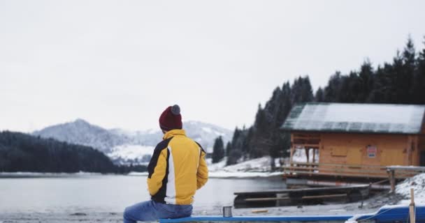 Посеред великого озера і поруч з дерев'яним будинком людина має час медитації, сидячи на блакитному човні і захоплюючись видом на озеро і ліс в зимовий день — стокове відео