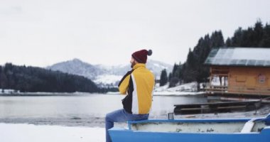 Büyük bir karlı göl ve orman karizmatik genç turist yanında o mavi bir tekne yanında oturan ve kameraya dönüm ve büyük gülümseyen görünümü hayran , turist turuncu bir ceket ve kırmızı giyiyor