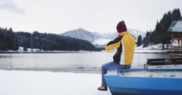 Hermoso turista de día de invierno sentado en el barco azul junto a la orilla del lago y admirar el paisaje de la naturaleza de montaña y bosque nevado, al lado del lago son una gran casa de madera — Vídeo de stock