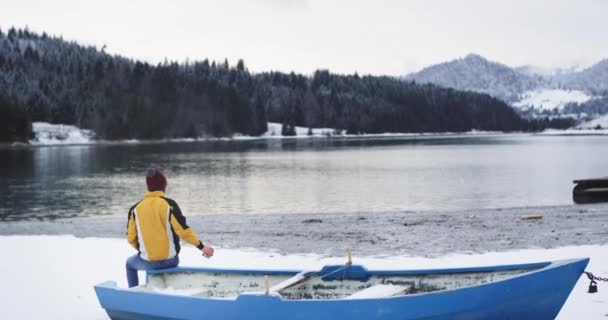 Big blue boat beside of a shore lake man with a orange jacket have a relaxing time admire all the view of a snowy forest and mountain drink some tea from iron cup — Stock Video