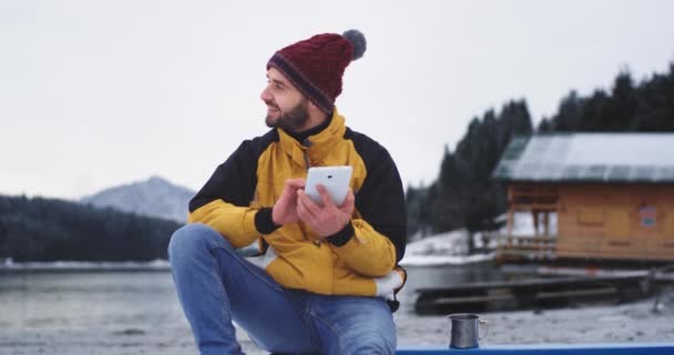 Jonge toerist aangekomen in een prachtig landschap met groot meer en bos, naast een houten huis heeft hij een kleine pauze chatten met een aantal vrienden of familieleden met behulp van een tablet, zeer gelukkig en — Stockvideo