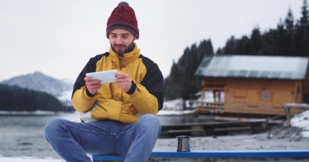 Lächelnder charismatischer junger Kerl, der mit einem Tablet Fotos von einem erstaunlichen Naturort mit Seewald und Bergen macht, er ist sehr begeistert von der Aussicht, im Hintergrund ein großes Holzhaus — Stockvideo