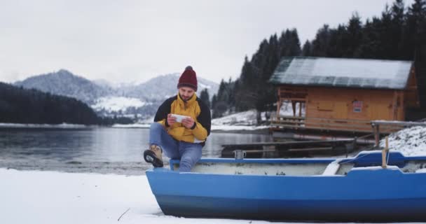 Красивый молодой турист отдохнуть он сидит на синей лодке рядом с берегом озера и смотрит через планшет, болтая, в удивительный зимний день — стоковое видео