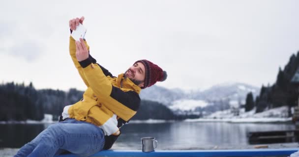 Зимний день турист прибыл к месту назначения удивительное место большого озера и снежного леса и горы он делает селфи с помощью планшета — стоковое видео