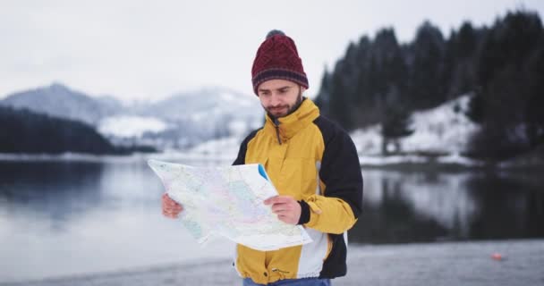 Joven turista en la chaqueta naranja comprobando en el mapa su ubicación, llegó a un hermoso lugar con un paisaje increíble de un gran lago y bosque nevado y montaña — Vídeos de Stock