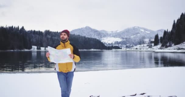 Giovane uomo con una mappa che controlla la sua posizione, cammina davanti alla telecamera intorno al lago riva e guardarsi intorno per trovare un posto per avere un po 'di pausa, bella vista sullo sfondo della montagna innevata — Video Stock