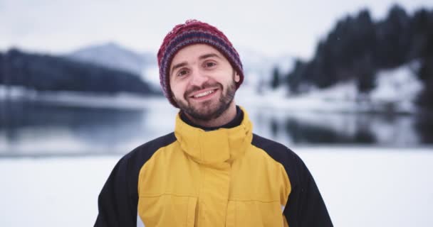 Большой улыбчивый молодой человек в красной шляпе посреди природы с красивым озером и заснеженной горой и лесом смотрит прямо в камеру и делает смешные лица — стоковое видео