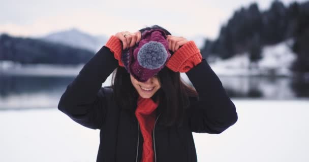 カメラにまっすぐ見て茶色の目の肖像画を持つカリスマ的な若い女性は、大きな湖と雪の森と山の素晴らしい背景を持つ自然の真ん中で彼女の赤い帽子をドレッシー — ストック動画