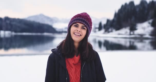 Momentos engraçados retrato na natureza em um dia de inverno uma linda jovem bonita com um grande sorriso, atrás de um grande lago e montanha olhando diretamente para a câmera e alguns amigos atirar a neve — Vídeo de Stock